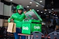 A delivery person wearing a green jacket and helmet holds a brown paper bag while standing beside a green insulated delivery bag with the logo 'Grab' on it. The setting appears to be inside a restaurant or food establishment with tables and chairs.