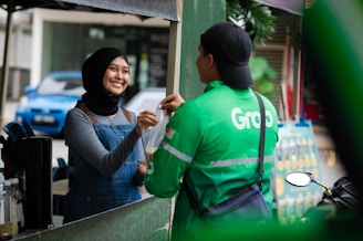 A woman wearing a hijab and a denim apron is smiling as she hands a plastic bag to a man wearing a green jacket with a logo. The background includes a parked car and some greenery.