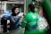 A woman wearing a hijab and a denim apron is smiling as she hands a plastic bag to a man wearing a green jacket with a logo. The background includes a parked car and some greenery.