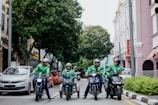 Four delivery riders wearing green jackets and helmets are on motorcycles on a city street. They are gathered in a lined formation in front of a row of parked cars. The street is flanked by greenery on one side and pastel-colored buildings on the other, creating a lively urban atmosphere.