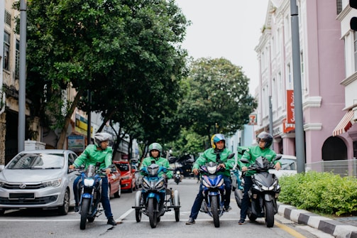 Four delivery riders wearing green jackets and helmets are on motorcycles on a city street. They are gathered in a lined formation in front of a row of parked cars. The street is flanked by greenery on one side and pastel-colored buildings on the other, creating a lively urban atmosphere.