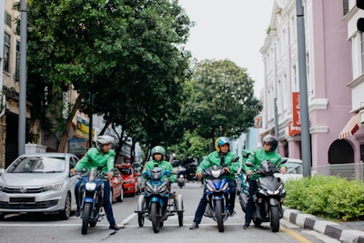 Four delivery riders wearing green jackets and helmets are on motorcycles on a city street. They are gathered in a lined formation in front of a row of parked cars. The street is flanked by greenery on one side and pastel-colored buildings on the other, creating a lively urban atmosphere.
