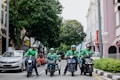 Four delivery riders wearing green jackets and helmets are on motorcycles on a city street. They are gathered in a lined formation in front of a row of parked cars. The street is flanked by greenery on one side and pastel-colored buildings on the other, creating a lively urban atmosphere.