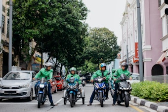 Four delivery riders wearing green jackets and helmets are on motorcycles on a city street. They are gathered in a lined formation in front of a row of parked cars. The street is flanked by greenery on one side and pastel-colored buildings on the other, creating a lively urban atmosphere.