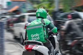 A GrabFood delivery person wearing a green uniform and helmet is riding a motorbike in a busy urban street amid traffic. The rider has a large insulated delivery bag on their back that features the GrabFood logo.