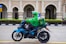 A delivery person wearing a green uniform and helmet rides a blue motorcycle. The motorcycle has a branded delivery box attached to the back. The scene includes an ornate building with arches and decorative columns in the background, suggesting an urban setting.