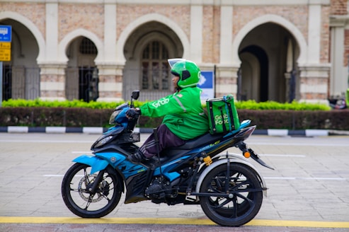 A delivery person wearing a green uniform and helmet rides a blue motorcycle. The motorcycle has a branded delivery box attached to the back. The scene includes an ornate building with arches and decorative columns in the background, suggesting an urban setting.