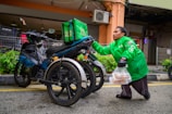 A person wearing a bright green jacket is holding a plastic bag with food containers, standing next to a three-wheeled motorcycle with a delivery box labeled 'Food' on it. The background shows a building exterior with metal fences, plants, and an air conditioning unit.