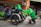 A person wearing a bright green jacket is holding a plastic bag with food containers, standing next to a three-wheeled motorcycle with a delivery box labeled 'Food' on it. The background shows a building exterior with metal fences, plants, and an air conditioning unit.