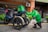 A person wearing a bright green jacket is holding a plastic bag with food containers, standing next to a three-wheeled motorcycle with a delivery box labeled 'Food' on it. The background shows a building exterior with metal fences, plants, and an air conditioning unit.