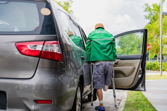 A person wearing a green Grab jacket is using crutches to get into a car. The car is parked by a curb, and there is greenery in the background. The person appears to have a prosthetic leg and is entering through the open rear door of the vehicle.