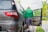A person wearing a green Grab jacket is using crutches to get into a car. The car is parked by a curb, and there is greenery in the background. The person appears to have a prosthetic leg and is entering through the open rear door of the vehicle.