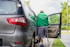 A person wearing a green Grab jacket is using crutches to get into a car. The car is parked by a curb, and there is greenery in the background. The person appears to have a prosthetic leg and is entering through the open rear door of the vehicle.