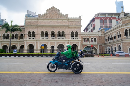 A person on a motorcycle rides past an ornate historical building featuring arches and intricate architectural details. The rider wears a green jacket and helmet, carrying a delivery box on the back of the motorcycle. The building is surrounded by a paved road and some palm trees, with modern skyscrapers visible in the background.