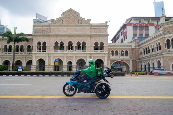 A person on a motorcycle rides past an ornate historical building featuring arches and intricate architectural details. The rider wears a green jacket and helmet, carrying a delivery box on the back of the motorcycle. The building is surrounded by a paved road and some palm trees, with modern skyscrapers visible in the background.