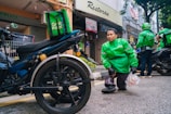 A person wearing a green Grab Food jacket is crouching near a motorcycle. The person is holding a plastic bag with food items. Another individual in a similar green jacket is in the background, also near a motorcycle. The setting appears to be a street in front of small shops.