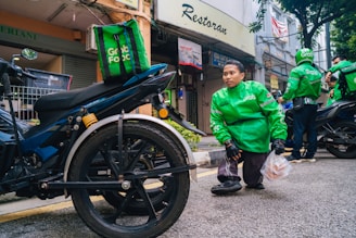 A person wearing a green Grab Food jacket is crouching near a motorcycle. The person is holding a plastic bag with food items. Another individual in a similar green jacket is in the background, also near a motorcycle. The setting appears to be a street in front of small shops.