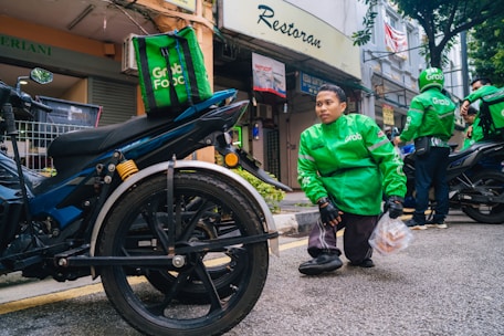 A person wearing a green Grab Food jacket is crouching near a motorcycle. The person is holding a plastic bag with food items. Another individual in a similar green jacket is in the background, also near a motorcycle. The setting appears to be a street in front of small shops.