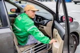 A person wearing a cap and a green jacket with the Grab logo is seated in a car, holding onto the door as it is open. The focus is on the interior and the person, with parts of the car like the dashboard, steering wheel, and side mirror visible.