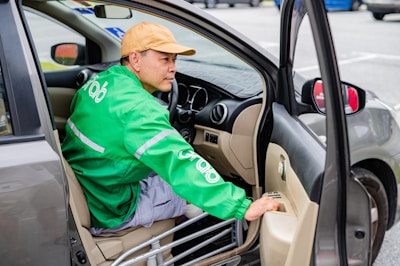 A person wearing a cap and a green jacket with the Grab logo is seated in a car, holding onto the door as it is open. The focus is on the interior and the person, with parts of the car like the dashboard, steering wheel, and side mirror visible.