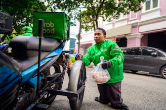A delivery person wearing a bright green GrabFood jacket is kneeling beside a motorcycle, holding a plastic bag with food items. The motorcycle is equipped with a green Grab insulated delivery bag. The person is on a city street with cars parked alongside and a building with white and pink walls in the background.