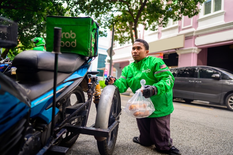 A delivery person wearing a bright green GrabFood jacket is kneeling beside a motorcycle, holding a plastic bag with food items. The motorcycle is equipped with a green Grab insulated delivery bag. The person is on a city street with cars parked alongside and a building with white and pink walls in the background.