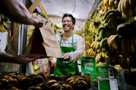 A man wearing a green apron is smiling and handing a GrabFood delivery bag to a customer in a fruit market. The market stall is filled with bunches of bananas hanging and displayed, with a prominent sign suggesting payment options through GrabPay.