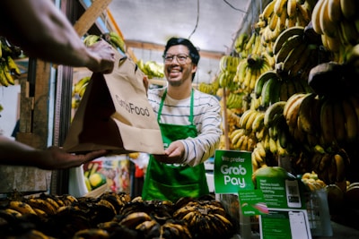 A man wearing a green apron is smiling and handing a GrabFood delivery bag to a customer in a fruit market. The market stall is filled with bunches of bananas hanging and displayed, with a prominent sign suggesting payment options through GrabPay.