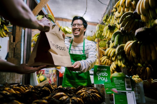 A man wearing a green apron is smiling and handing a GrabFood delivery bag to a customer in a fruit market. The market stall is filled with bunches of bananas hanging and displayed, with a prominent sign suggesting payment options through GrabPay.