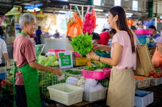 A vendor and a customer exchange fresh green vegetables at a market stall. The vendor, wearing a green apron, stands behind a counter filled with assorted produce, including baskets of leafy greens and other vegetables. The customer, smiling and carrying a reusable shopping bag, is engaging in conversation. The background shows a bustling market environment with other shoppers and stalls filled with colorful goods.
