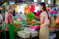 A vendor and a customer exchange fresh green vegetables at a market stall. The vendor, wearing a green apron, stands behind a counter filled with assorted produce, including baskets of leafy greens and other vegetables. The customer, smiling and carrying a reusable shopping bag, is engaging in conversation. The background shows a bustling market environment with other shoppers and stalls filled with colorful goods.