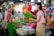 A vendor and a customer exchange fresh green vegetables at a market stall. The vendor, wearing a green apron, stands behind a counter filled with assorted produce, including baskets of leafy greens and other vegetables. The customer, smiling and carrying a reusable shopping bag, is engaging in conversation. The background shows a bustling market environment with other shoppers and stalls filled with colorful goods.