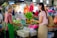 A vendor and a customer exchange fresh green vegetables at a market stall. The vendor, wearing a green apron, stands behind a counter filled with assorted produce, including baskets of leafy greens and other vegetables. The customer, smiling and carrying a reusable shopping bag, is engaging in conversation. The background shows a bustling market environment with other shoppers and stalls filled with colorful goods.