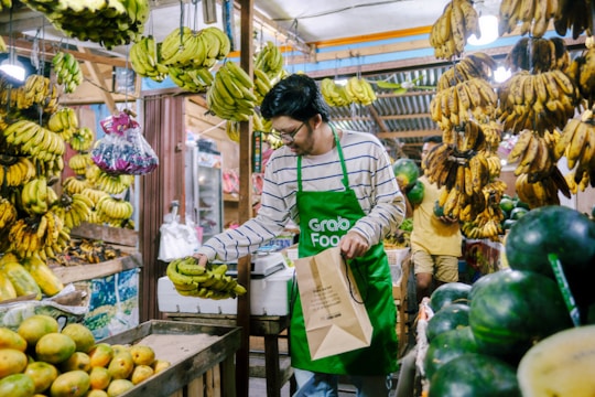 A person wearing a green apron is selecting bananas in a vibrant market filled with hanging banana bunches. There are fresh fruits like bananas, watermelons, and mangoes arranged around the stall, creating a lively and bustling atmosphere.