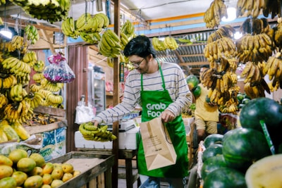 A person wearing a green apron is selecting bananas in a vibrant market filled with hanging banana bunches. There are fresh fruits like bananas, watermelons, and mangoes arranged around the stall, creating a lively and bustling atmosphere.