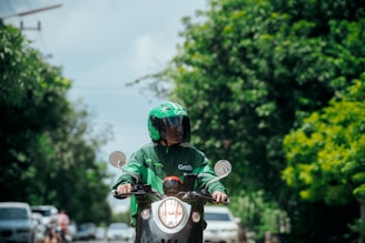 A person wearing a green Grab uniform and helmet is riding a motorcycle. The background includes lush green trees and a few vehicles on the road.