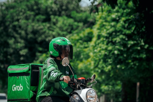 A delivery person wearing a green helmet and jacket rides a scooter. The scooter features a large green delivery box on the back, and the surroundings include lush green trees, suggesting an outdoor environment.
