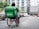 A person in a hoodie, using a wheelchair equipped with a large green delivery backpack labeled 'Grab.' They are on a paved street with parked cars on the side and a multi-story residential building in the background.