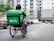 A person in a hoodie, using a wheelchair equipped with a large green delivery backpack labeled 'Grab.' They are on a paved street with parked cars on the side and a multi-story residential building in the background.