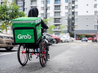 A person in a hoodie, using a wheelchair equipped with a large green delivery backpack labeled 'Grab.' They are on a paved street with parked cars on the side and a multi-story residential building in the background.