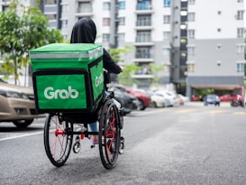A person in a hoodie, using a wheelchair equipped with a large green delivery backpack labeled 'Grab.' They are on a paved street with parked cars on the side and a multi-story residential building in the background.