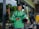 A delivery rider wearing a green Grab uniform stands outside a glass-fronted shop. He holds a helmet in one hand and a smartphone in the other. His green backpack is slung over his shoulders, and he appears focused on the phone. The background includes a glimpse of greenery and a restaurant interior.