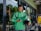 A delivery rider wearing a green Grab uniform stands outside a glass-fronted shop. He holds a helmet in one hand and a smartphone in the other. His green backpack is slung over his shoulders, and he appears focused on the phone. The background includes a glimpse of greenery and a restaurant interior.