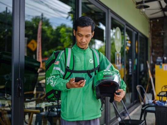 A delivery rider wearing a green Grab uniform stands outside a glass-fronted shop. He holds a helmet in one hand and a smartphone in the other. His green backpack is slung over his shoulders, and he appears focused on the phone. The background includes a glimpse of greenery and a restaurant interior.