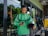 A delivery rider wearing a green Grab uniform stands outside a glass-fronted shop. He holds a helmet in one hand and a smartphone in the other. His green backpack is slung over his shoulders, and he appears focused on the phone. The background includes a glimpse of greenery and a restaurant interior.