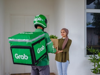 A delivery person wearing Grab-branded attire, including a green helmet and jacket, hands over a brown paper bag to a smiling young woman standing at a door. The woman is dressed casually with a headscarf and there are green plants near the entrance.