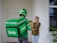 A delivery person wearing Grab-branded attire, including a green helmet and jacket, hands over a brown paper bag to a smiling young woman standing at a door. The woman is dressed casually with a headscarf and there are green plants near the entrance.