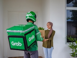 A delivery person wearing Grab-branded attire, including a green helmet and jacket, hands over a brown paper bag to a smiling young woman standing at a door. The woman is dressed casually with a headscarf and there are green plants near the entrance.