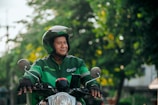 A person wearing a helmet and a green jacket with a Grab logo is riding a motorcycle. The background features a blurred mix of greenery and urban elements, suggesting an outdoor setting.