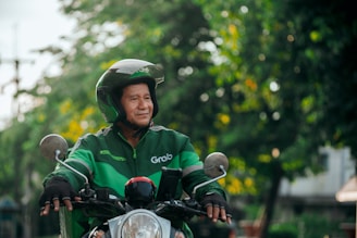 A person wearing a helmet and a green jacket with a Grab logo is riding a motorcycle. The background features a blurred mix of greenery and urban elements, suggesting an outdoor setting.